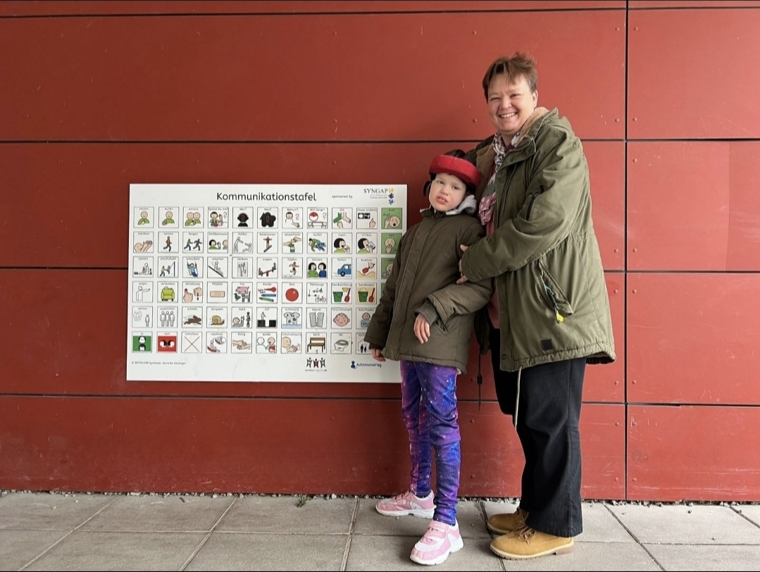 Spielplatztafel mit METACOM Symbolen an roter Wand, rechts daneben Mama mit Kind